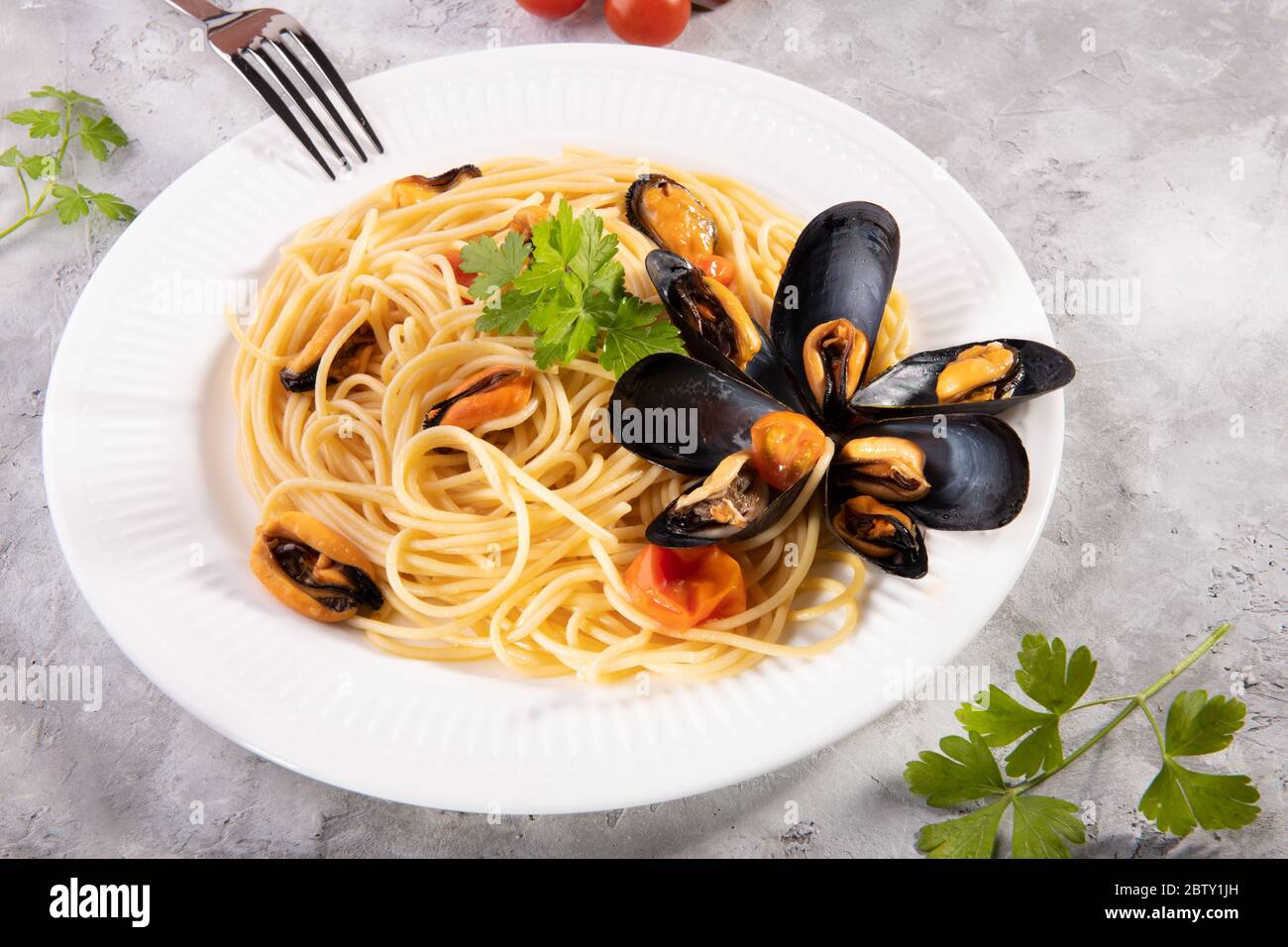 Spaghetti de pâtes italiennes typiques avec moules et tomates sur une assiette blanche avec ingrédients sur fond gris Banque D'Images