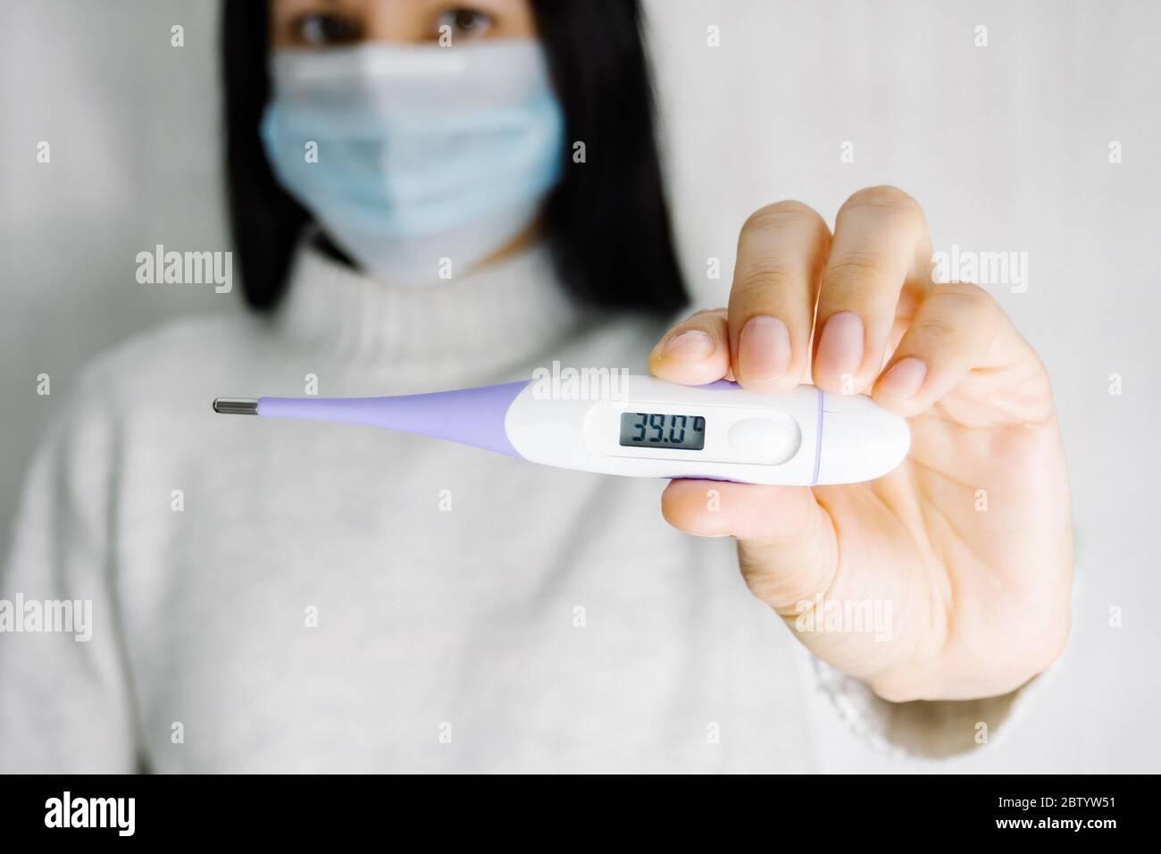 Jeune femme caucasienne en masque chirurgical tenant un thermomètre électronique dans les mains. Température élevée à l'écran, mise au point sélective Banque D'Images