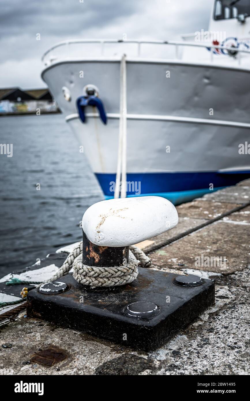 Borne blanche sur un quai avec des amarres serrées entre elle et un bateau à coque blanc et bleu. Banque D'Images