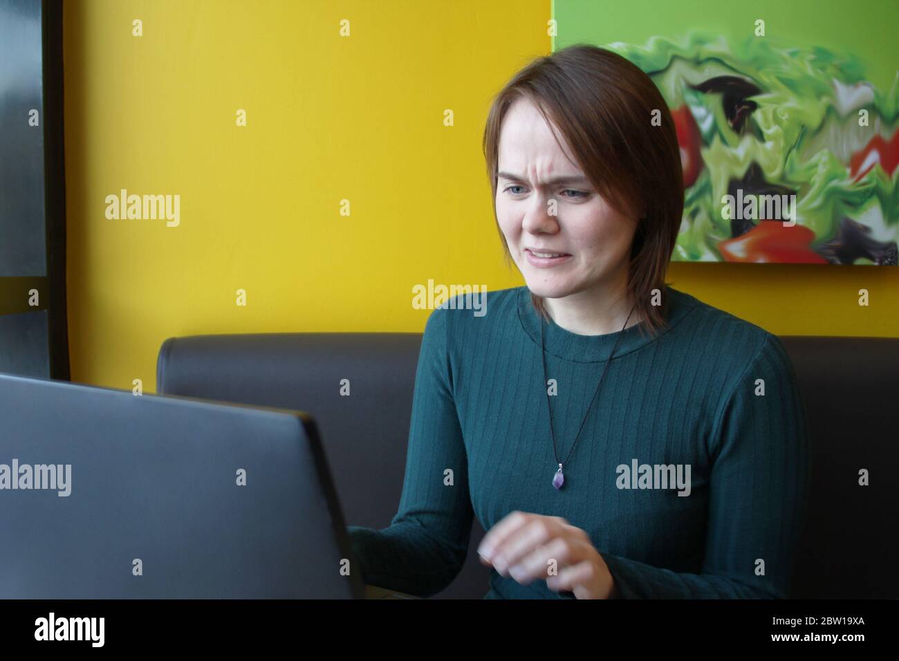 Une fille s'assoit, travaille à un ordinateur portable à une table dans un café sur un canapé marron contre un mur jaune. Au visage, on trouve une expression d'hostilité, de dégoût, d'abomination, de tension. Banque D'Images