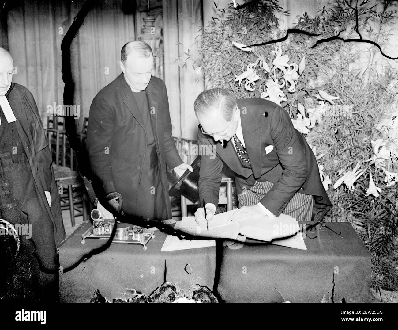 M. Joseph P Kennedy, ambassadeur américain, Dévoilée dans la cathédrale de Winchester, une vitrail qui a été donné à la cathédrale par les Américains en hommage à la vie et au caractère du roi George V. l'une des lumières contient une représentation de la figure à genoux du roi portant les robes de l'ordre de Le Garter'. La fenêtre est le travail de M. Hugh Easton. Photos, M. Joseph P Kennedy signant le livre des personnalités de la cathédrale. Il est surveillé par le doyen de Winchester. 12 juillet 1938 Banque D'Images