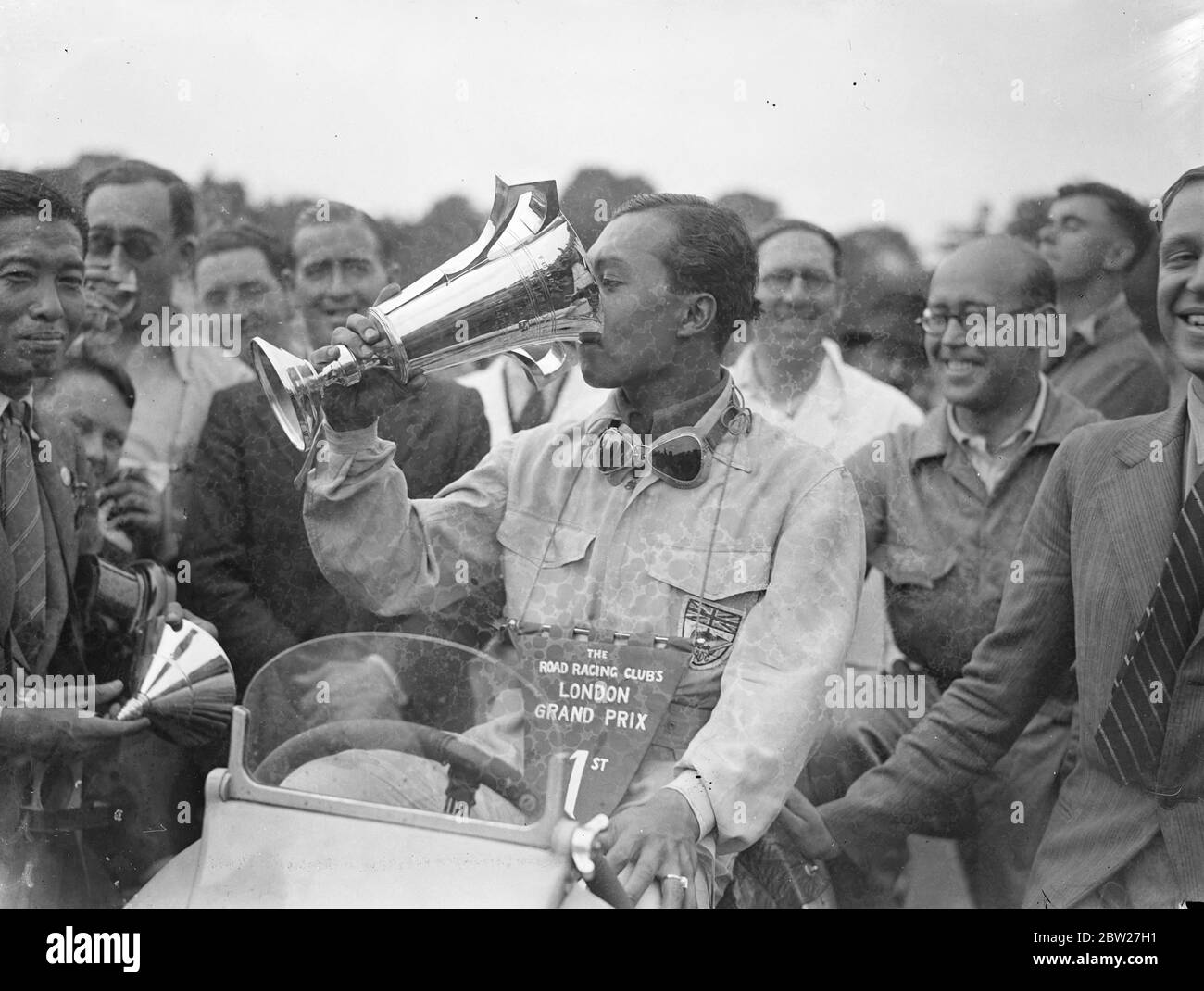 Le Prince Birabongse de Siam [Prince Bira de Siam / Thaïlande], pilotant une ERA [automobiles anglaises] a remporté le premier Grand Prix de Londres sur le nouveau circuit de course de Crystal Palace. I. F. Cornell (ERA) est deuxième et P. Maclure (Riley) troisième. Le prince Birabongse buvant de la coupe après sa victoire. 17 juillet 1937 Banque D'Images