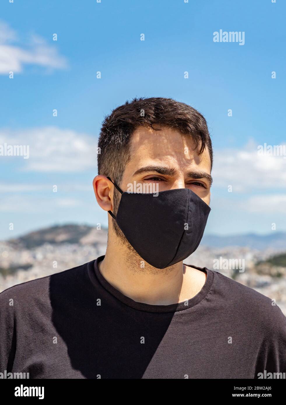 Athènes Grèce coronavirus Days. Jeune homme portant un masque de protection, un paysage urbain d'Athènes et un fond bleu ciel. Banque D'Images