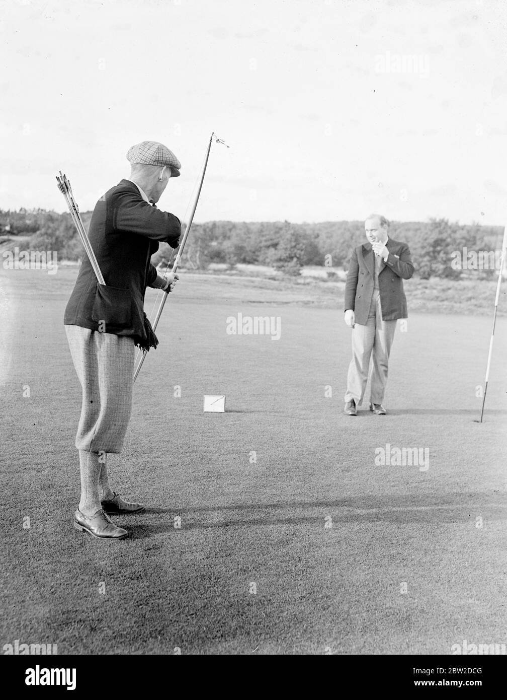 Jouant avec Bow et Arrow, le major J G Hayter, expert reconnu dans le sport annuel du golf Bow et Arrow, a rencontré le colonel E St George Kirke, le capitaine du club, dans un match au Hankley Common Golf Club, Surrey. Il a concédé un coup un gale. Le major Hayter tire sa flèche dans une boîte au lieu d'un trou. Sa moyenne pour la plupart des cours de Surrey est dans les années 70. Expositions de photos : le Major J G Hayter, qui tourne dans la boîte, est regardé par son adversaire, le Col E St George Kirke. 18 octobre 1938 Banque D'Images