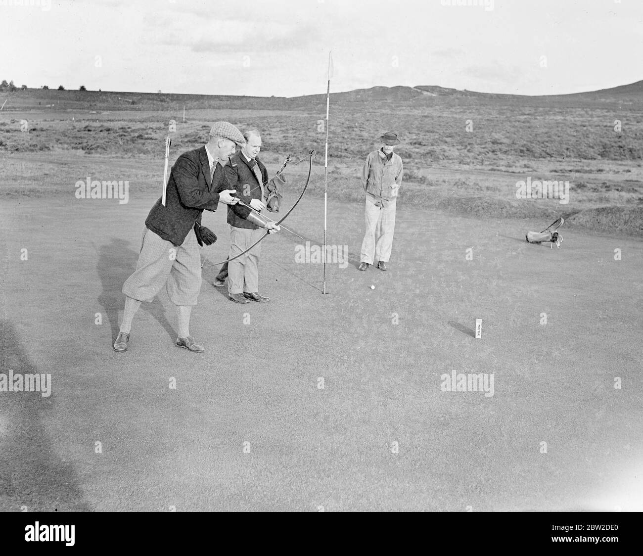 Jouant avec Bow et Arrow, le major J G Hayter, expert reconnu dans le sport annuel du golf Bow et Arrow, a rencontré le colonel E St George Kirke, le capitaine du club, dans un match au Hankley Common Golf Club, Surrey. Il a concédé un coup un gale. Le major Hayter tire sa flèche dans une boîte au lieu d'un trou. Sa moyenne pour la plupart des cours de Surrey est dans les années 70. Expositions de photos : le Major J G Hayter, qui tourne dans la boîte, est regardé par son adversaire, le Col E St George Kirke. 18 octobre 1938 Banque D'Images