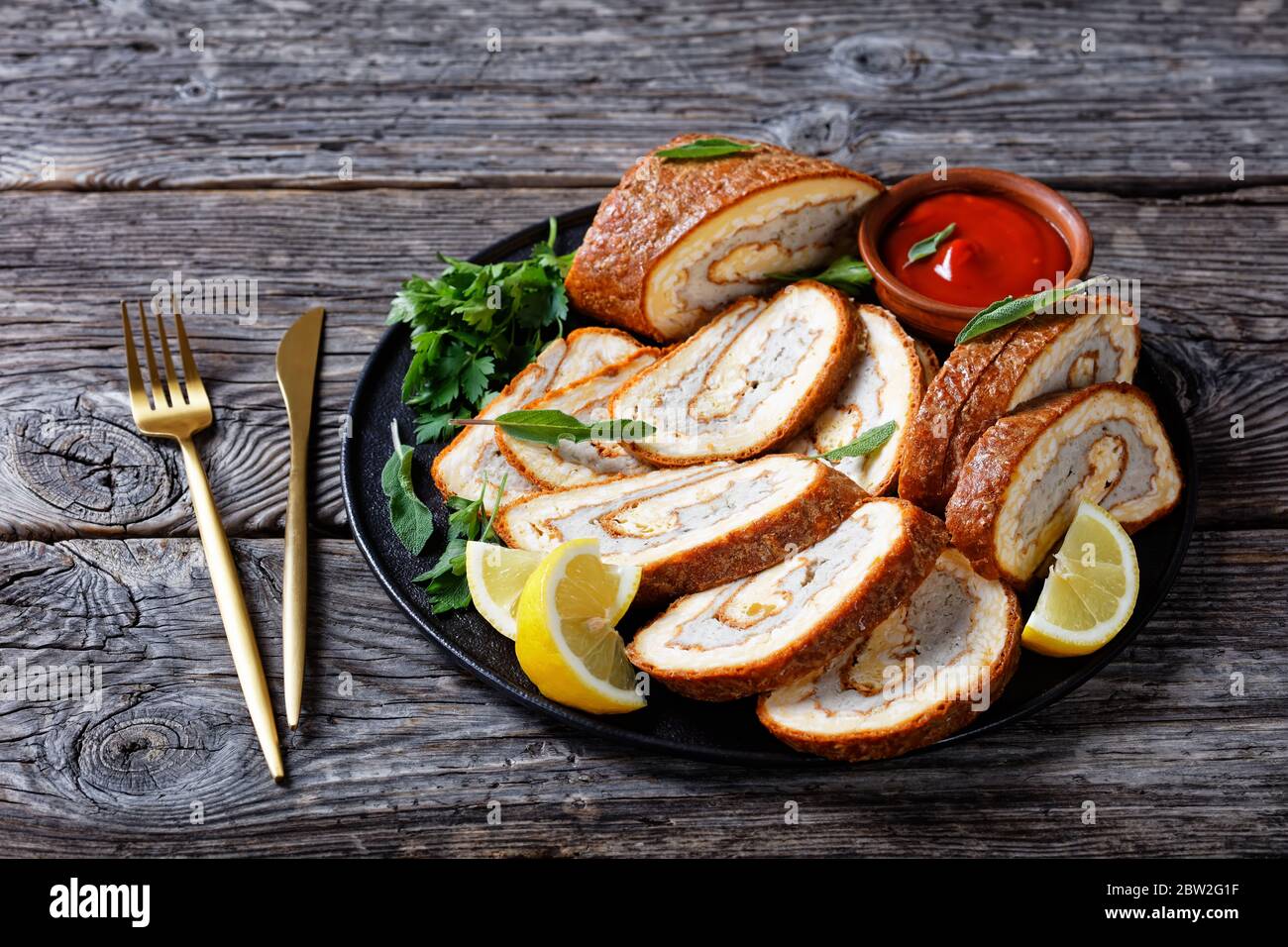 Omelette sur une assiette noire avec citron, ketchup et persil, rouler avec garniture de viande hachée sur une table en bois gris avec fourchette et couteau dorés, chapeau de terre Banque D'Images