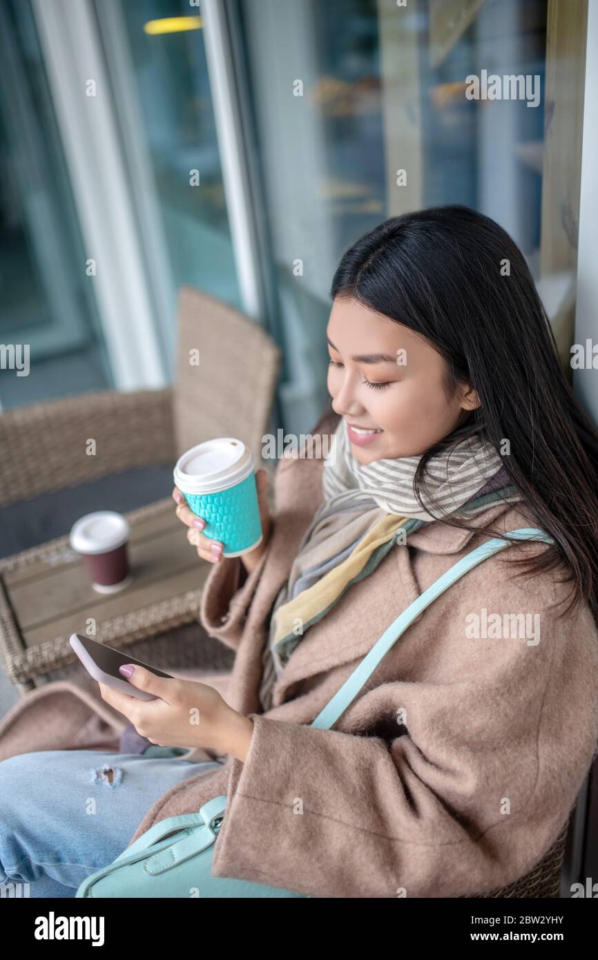 Brunette femme en beige manteau assis, boire du café, vérifier son téléphone portable Banque D'Images
