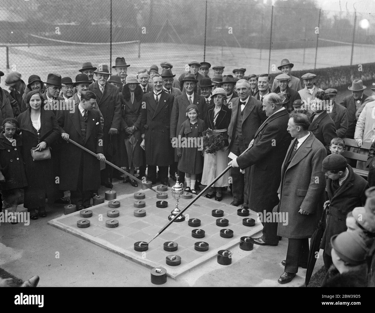Première séance ouverte - projet d'air sur table de six pieds carrés , ouverte dans un parc de Londres . Le premier tournoi ouvert - air drafts à jouer à Londres a été commencé à Downhills Park , Londres , quand les concurrents ont joué sur une table de six pieds carrés . Les pièces ou les hommes ont six pouces de diamètre et sont déplacés par les hommes avec cinq pieds cendre bâtons . Photos ; M. Cohen , le champion britannique ( à gauche ) et M. Keysen ( à droite ) , jouant le premier match du tournoi , assisté par le conseiller Morley et sa femme qui a ouvert le match . 28 avril 1934 Banque D'Images