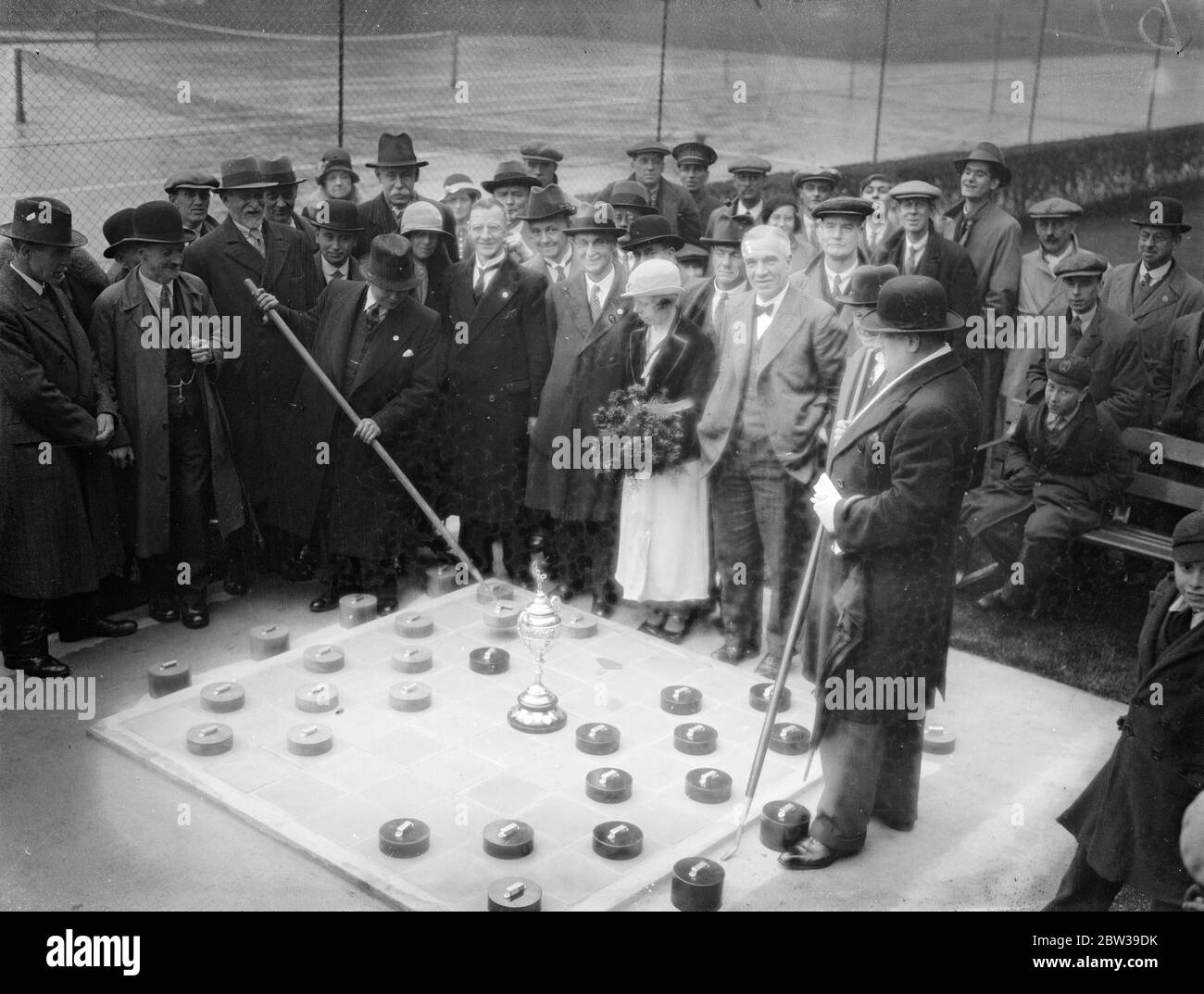 Première séance ouverte - projet d'air sur table de six pieds carrés , ouverte dans un parc de Londres . Le premier tournoi ouvert - air drafts à jouer à Londres a été commencé à Downhills Park , Londres , quand les concurrents ont joué sur une table de six pieds carrés . Les pièces ou les hommes ont six pouces de diamètre et sont déplacés par les hommes avec cinq pieds cendre bâtons . Photos ; M. Cohen , le champion britannique ( à gauche ) et M. Keysen ( à droite ) , jouant le premier match du tournoi , assisté par le conseiller Morley et sa femme qui a ouvert le match . 28 avril 1934 Banque D'Images