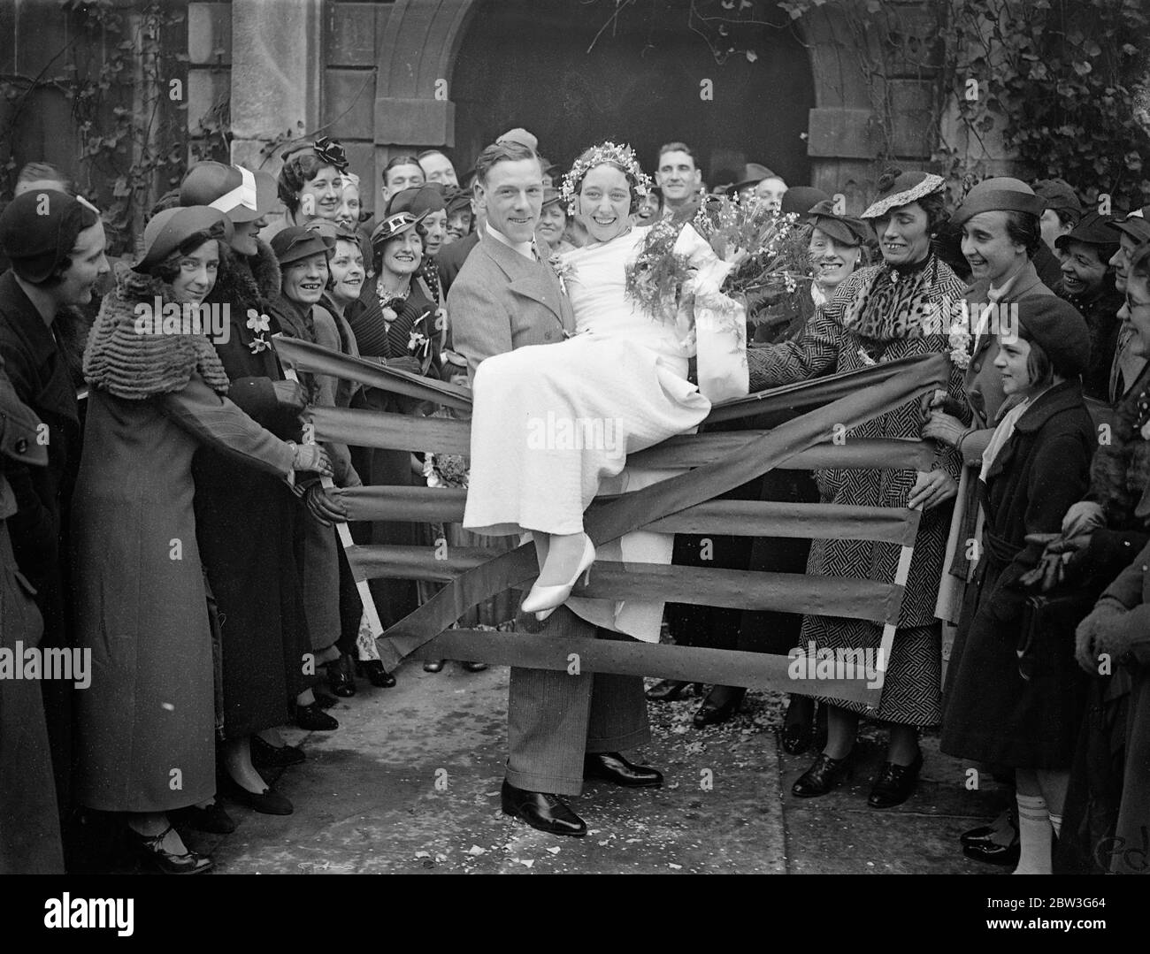 Le marié lève l'athlète mariée sur l'obstacle après , après le mariage à St Clément Danes . Mlle Lily Smith , coureur de fond des Olympiades de Londres , a été soulevée par son époux Thomas William Clements , lorsqu'elle était mariée à l'église St Clément Danes , Strand . La robe et la coiffure de la mariée ' s Lily ' de nénuphars de la vallée ont été faites par Mlle JO Parsons , le capitaine de la campagne. Photos montre , le marié levant sa mariée d'athlète sur l'obstacle après la cérémonie . 11 avril 1936 Banque D'Images