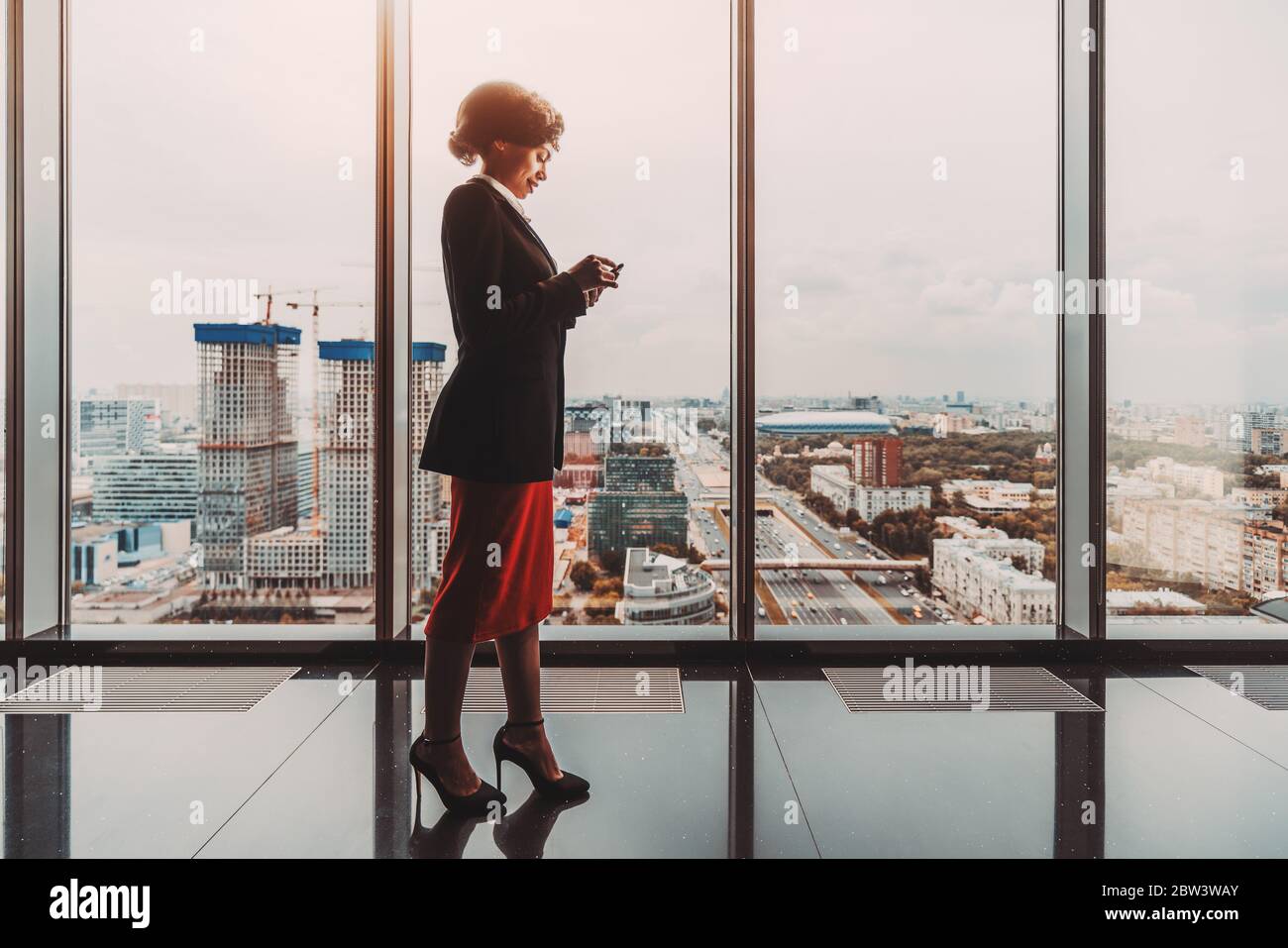 Silhouette d'une charmante femme d'affaires afro-américaine joyeuse utilisant son smartphone pour répondre à un message en se tenant devant une fenêtre panoramique Banque D'Images