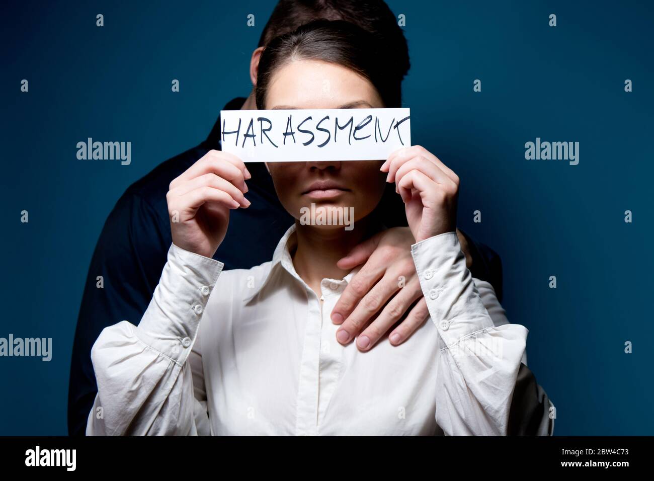 Une jeune femme en vêtements de bureau ferme les yeux avec un morceau de papier avec les mots harcèlement, un homme se tient derrière elle Banque D'Images