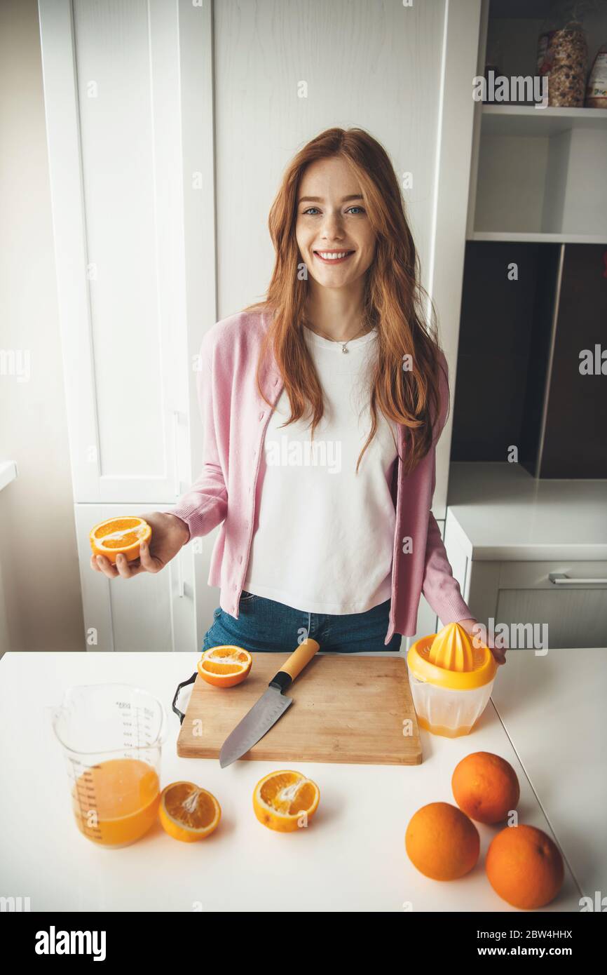 Une femme caucasienne aux cheveux rouges avec des taches de rousseur fait du jus d'orange dans la cuisine tout en souriant en piquant à l'appareil photo Banque D'Images
