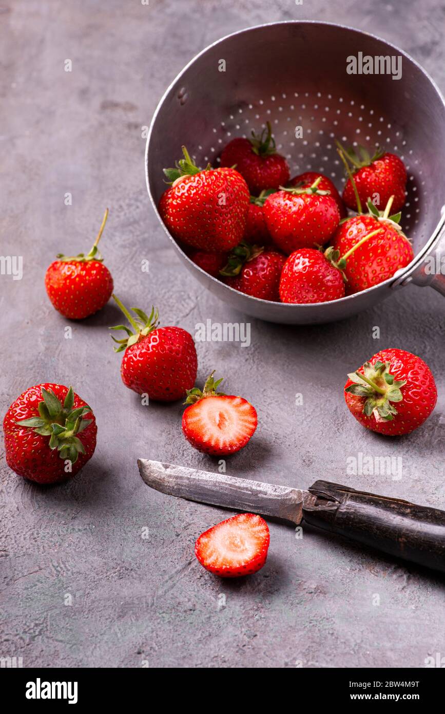 Sur fond gris texturé, fraises mûres dans une vieille passoire et quelques tranches avec un couteau vintage. Banque D'Images