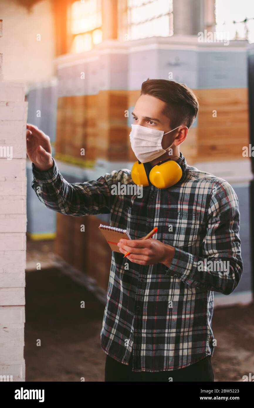 Un superviseur professionnel dans un masque médical et un casque de protection inspectant les stocks de bois. Responsable, contrôle des employés d'entrepôt, c Banque D'Images