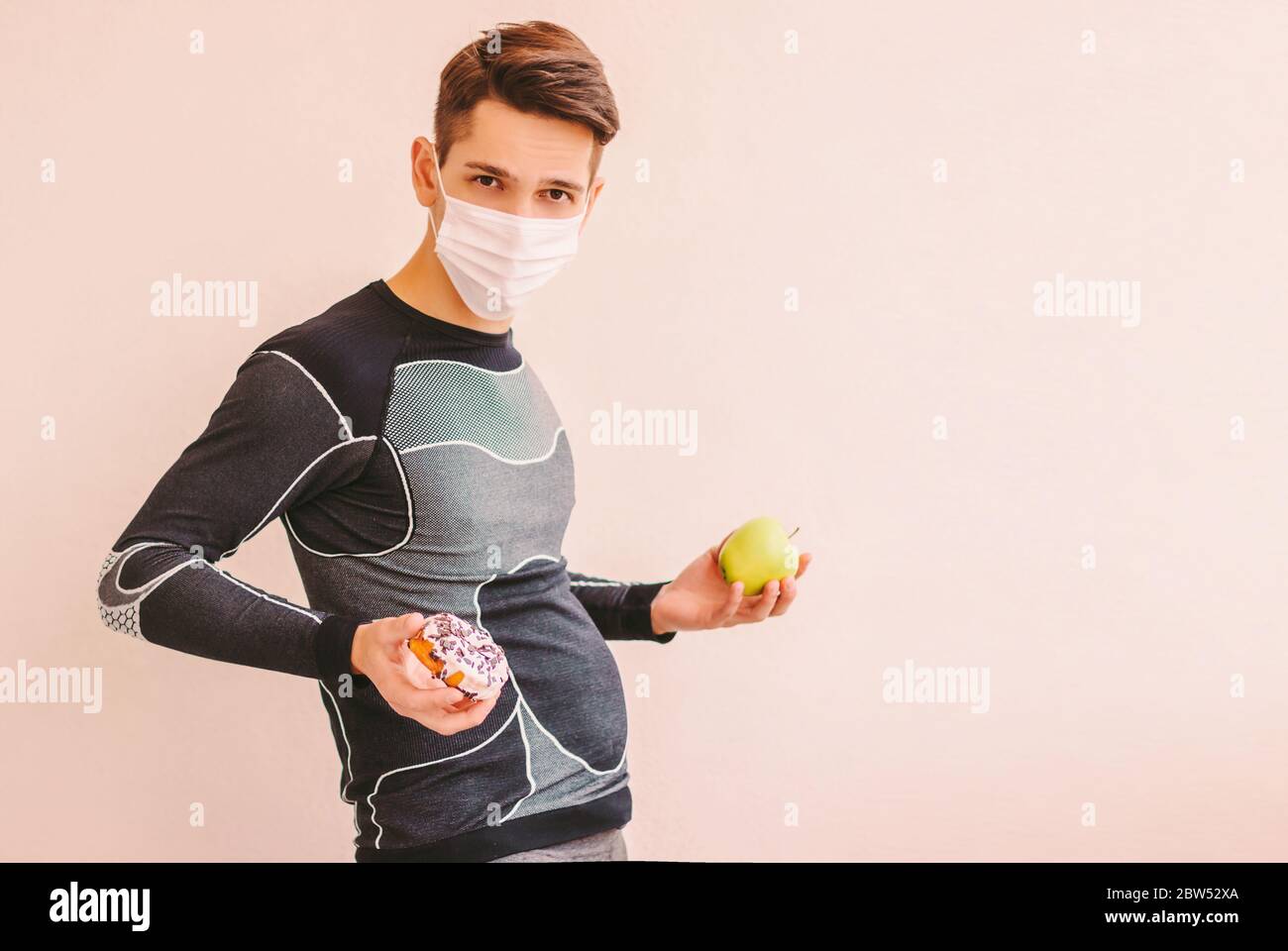 Jeune homme de sport gras dans le masque protecteur tient la pomme fraîche et le donut doux dans les mains. Drôle d'homme de forme physique dans le masque médical avec le grand ventre choisir betwee Banque D'Images