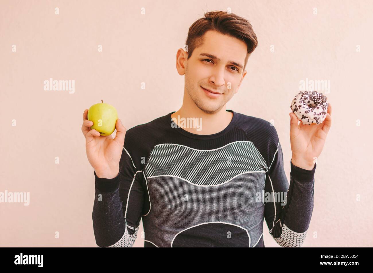 Portrait de jeunes sportifs heureux tenant un en-cas de beignet doux et une pomme fraîche dans les mains. Jeune homme de forme physique incertain choisir entre des fruits sains et Banque D'Images