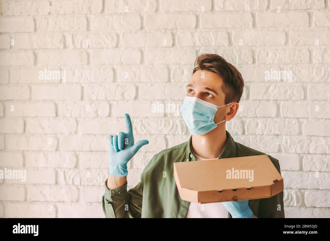 Happy hippster Guy courier dans le masque médical et les gants en caoutchouc tiennent la boîte à pizza en carton dans la main, regardez, pointez vers l'espace de copie. Service de livraison de l'homme Banque D'Images