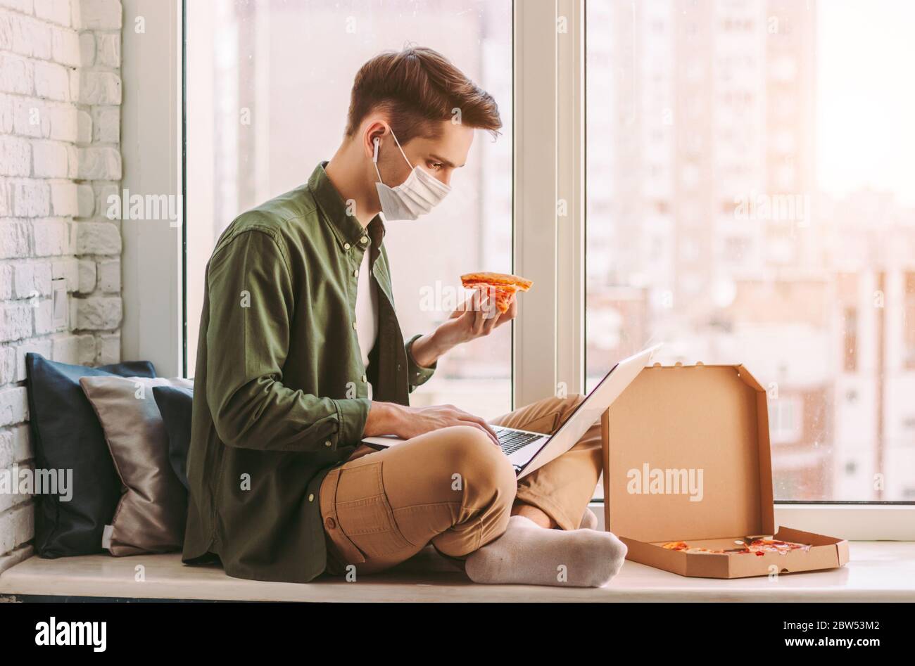Homme d'affaires affamé dans le masque médical manger pizza snack, utiliser l'ordinateur portable pour le travail à domicile. Un homme hipster élégant dans un masque de protection pour une tranche de pizza Banque D'Images