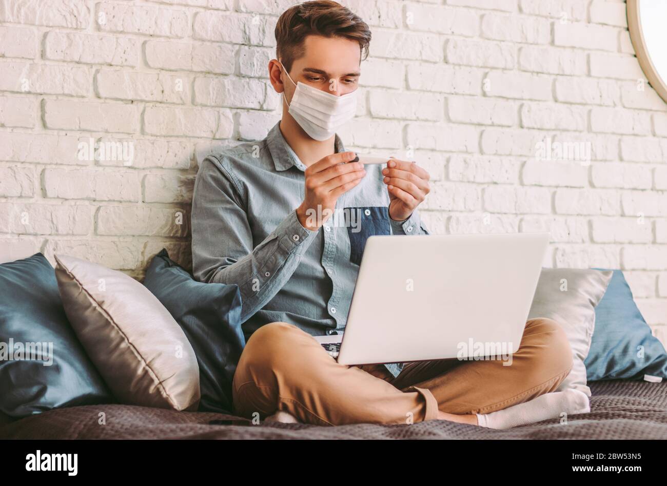 Malade hipster homme étudiant dans le masque médical de visage regarder le thermomètre numérique dans les mains, le travail à la maison. Jeune homme d'affaires indépendant dans l'utilisation de masque de protection Banque D'Images