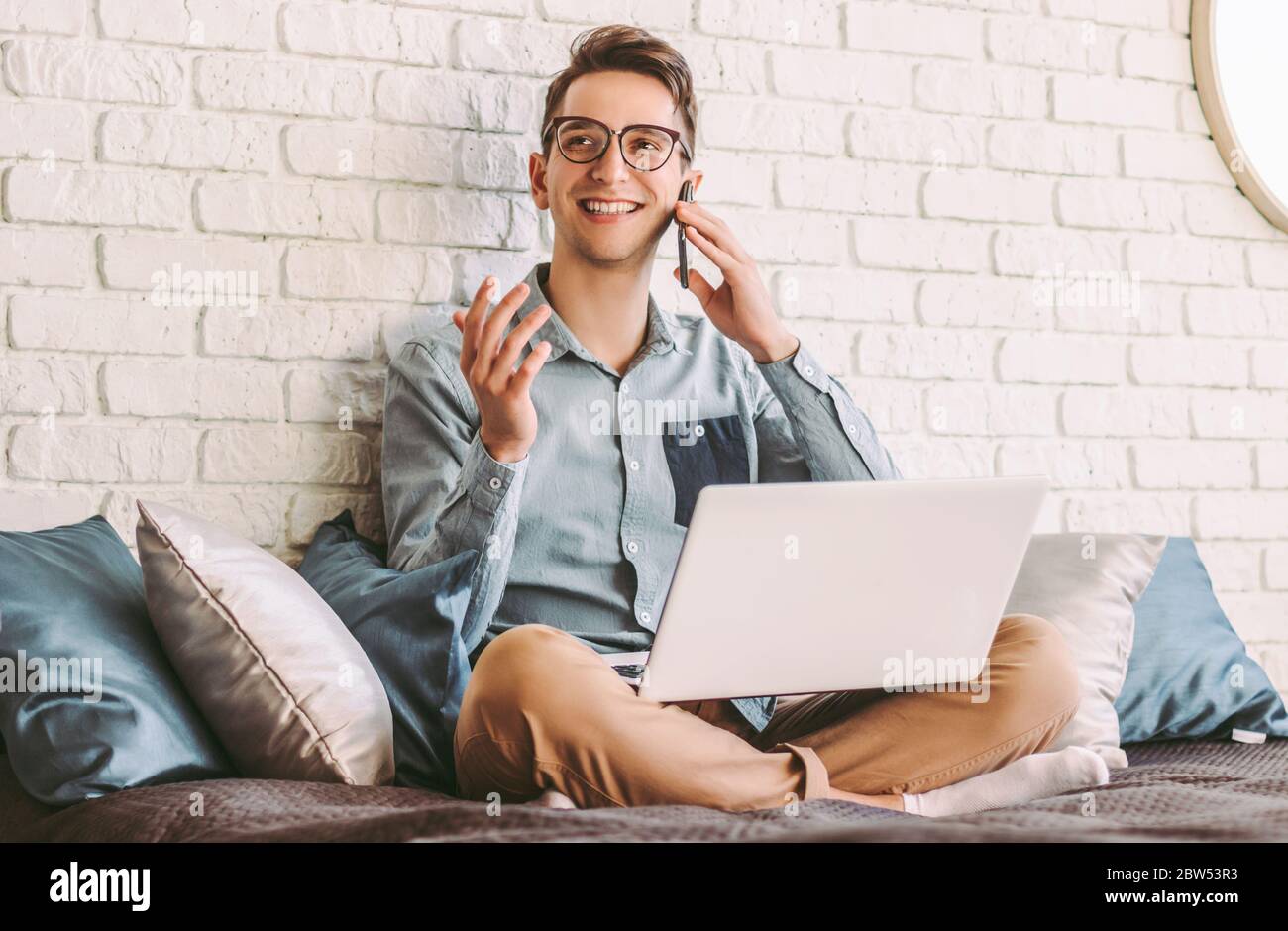 Un jeune homme d'affaires heureux dans des lunettes parlant téléphone mobile et souriant tout en travaillant sur ordinateur portable au bureau à domicile. Occupé chic hippster homme étudiant ont le pH de cellule Banque D'Images