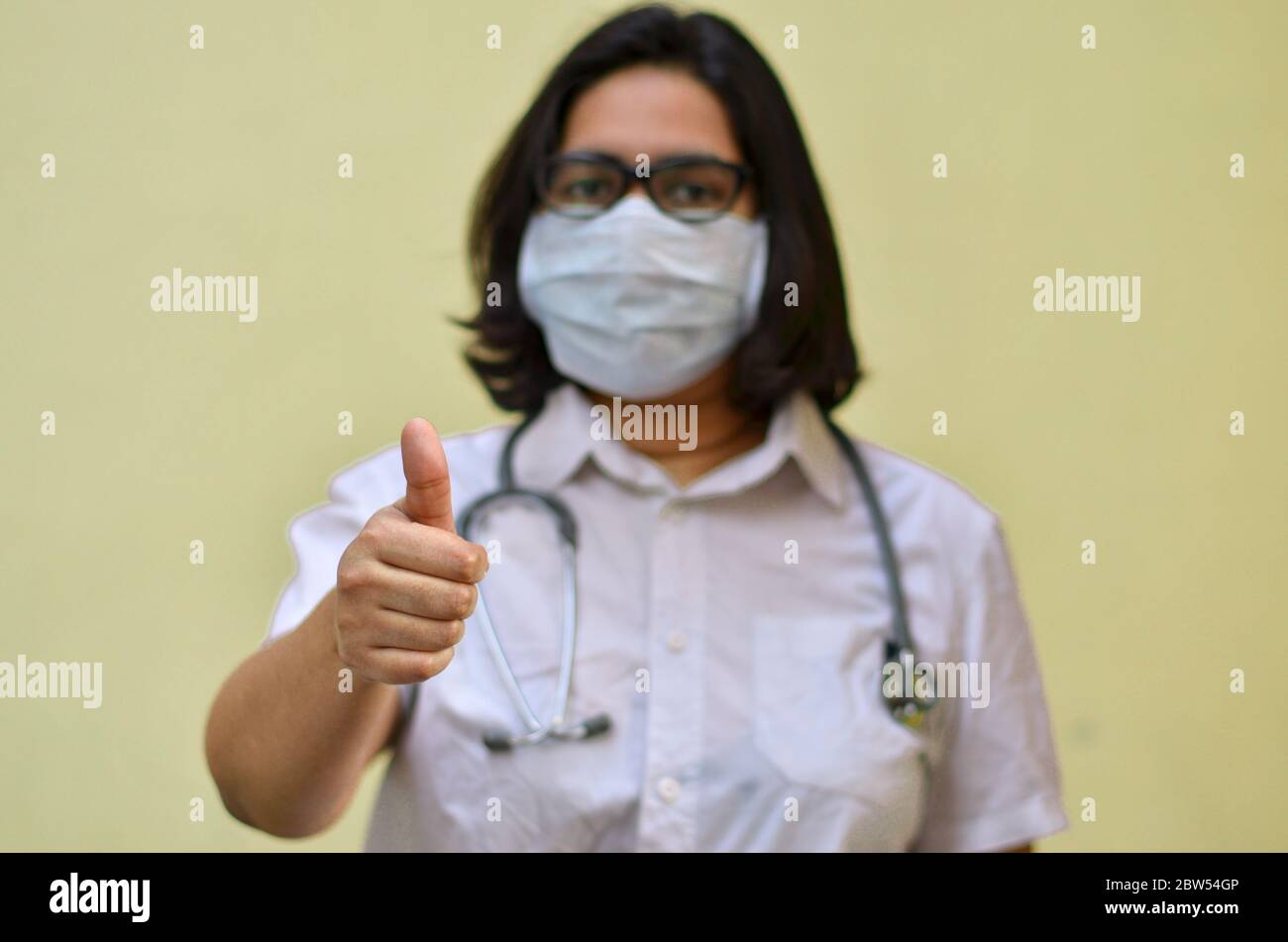 Portrait d'une jeune travailleuse médicale montrant un signe de pouce vers le haut avec sa main droite portant un masque chirurgical pour se protéger de Corona V. Banque D'Images