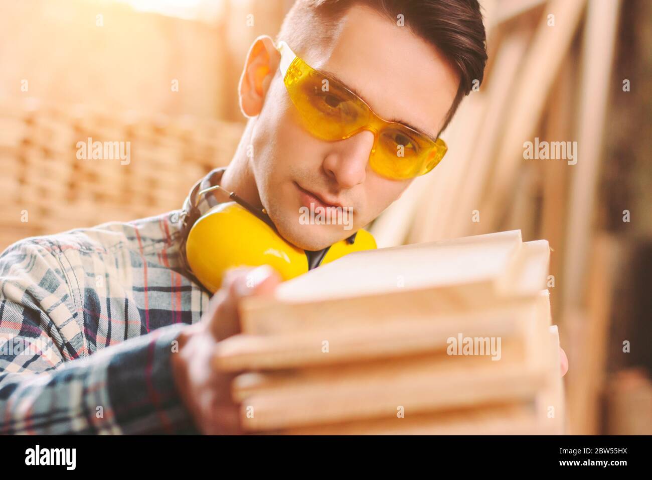 Gros plan d'un fabricant d'armoires sûr en lunettes de protection et écouteurs tenant une pile de matériaux en bois dans les mains. Jeune menuisier de sexe masculin travaillant à la scie Banque D'Images
