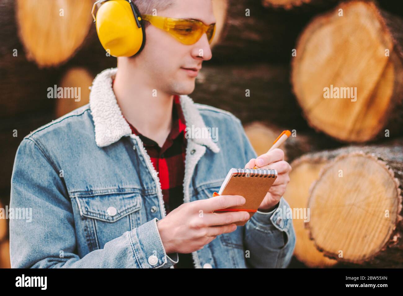 Portrait d'un jeune ouvrier d'entrepôt de bois d'œuvre dans des lunettes de protection et des écouteurs inspectant les troncs d'arbre coupés. Homme Forester dans la veste denim vérifiant scimi Banque D'Images