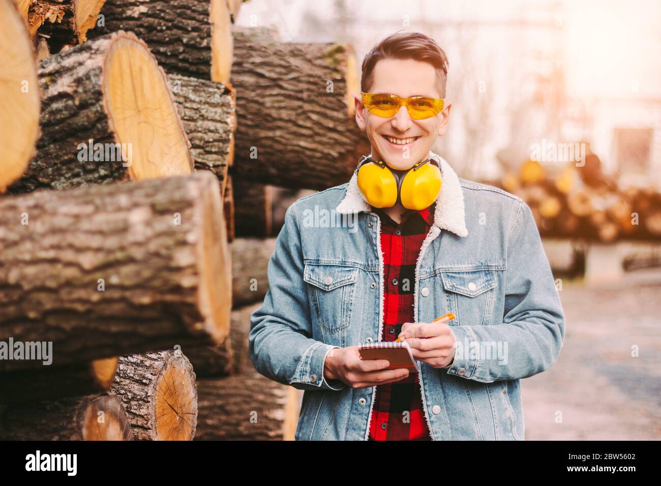 Bonne forestier dans les lunettes de protection et les casques inspectant l'inventaire d'entrepôt de bois d'œuvre. Coupe-bois jeune homme en veste denim comptant la bûche de l'arbre coupée Banque D'Images