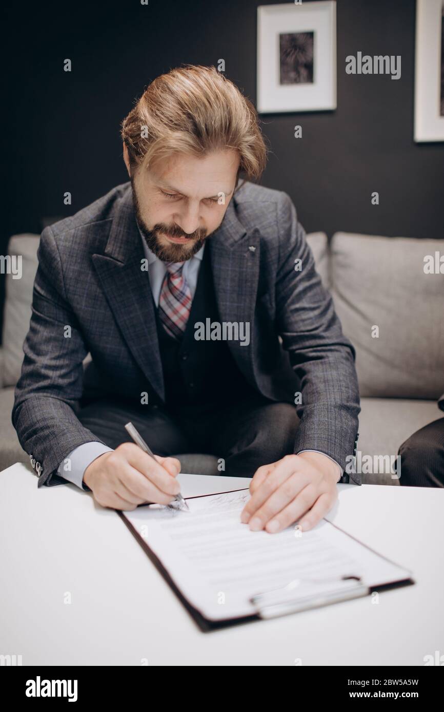 Homme à barbe mûr vêché de vêtements formels signant des documents tout en étant assis sur un canapé gris. Homme d'affaires compétent qui commence une relation financière avec une nouvelle société Banque D'Images