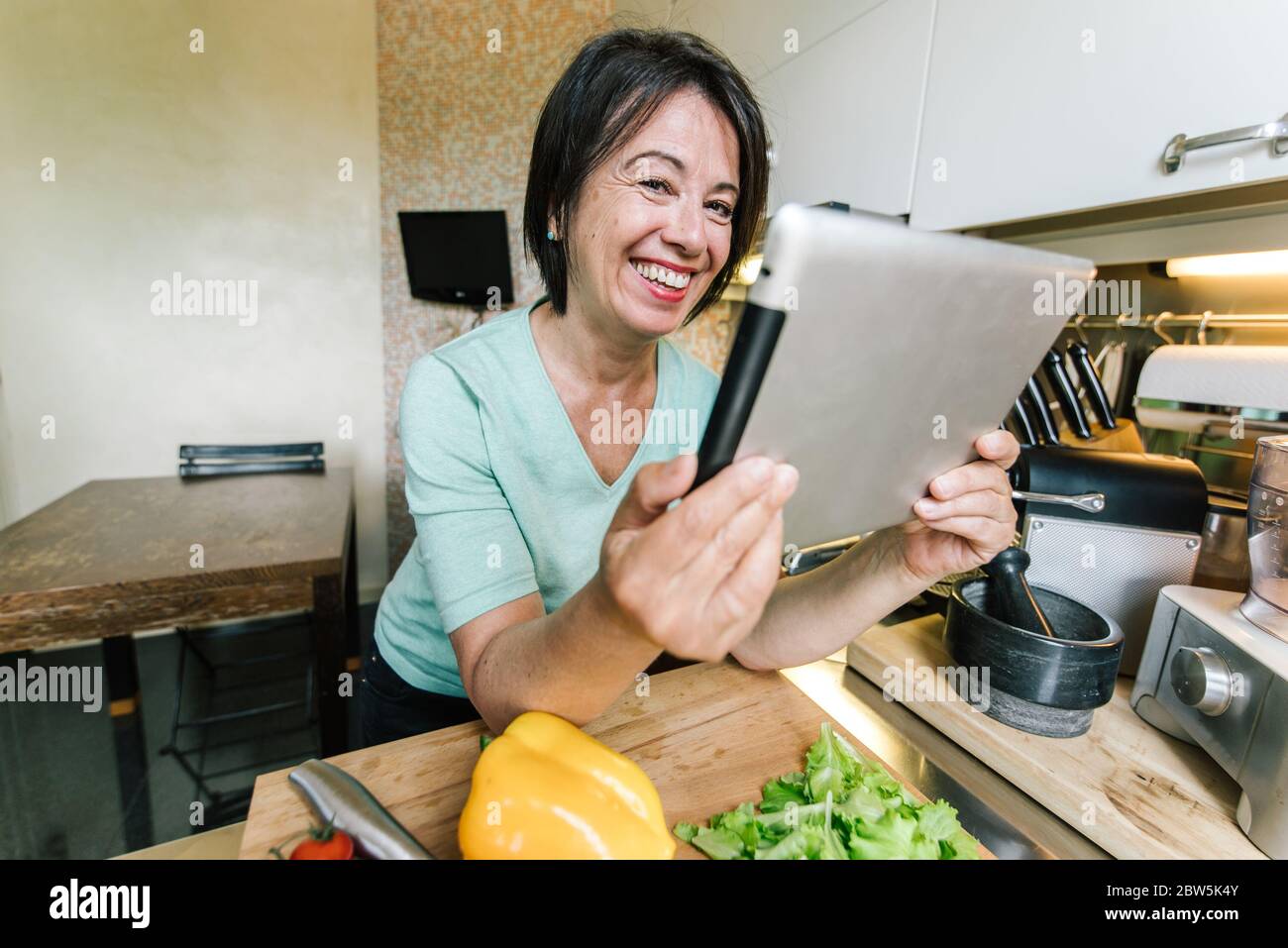 Femme aîâgée chef cuisinier dans la cuisine à la maison lisant la recette sur un appareil de tablette. Banque D'Images