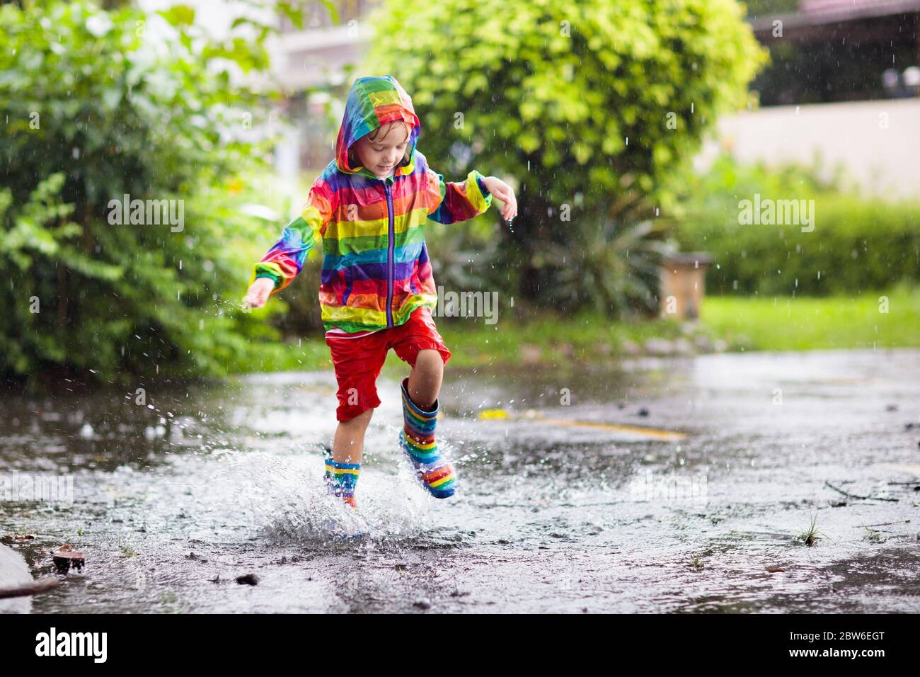 Enfant jouant dans la pluie dans le parc d'automne. Enfant sautant dans un  sol boueux le jour de l'automne pluvieux. Petit garçon en bottes de pluie  et veste arc-en-ciel dehors en lourd