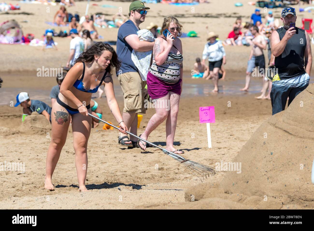 New River Beach, Nouveau-Brunswick, Canada - le 7 juillet 2018 : le concours annuel de sculpture sur sable. Une femme tatouée ramasse du sable à côté d'une sculpture de sable. Banque D'Images