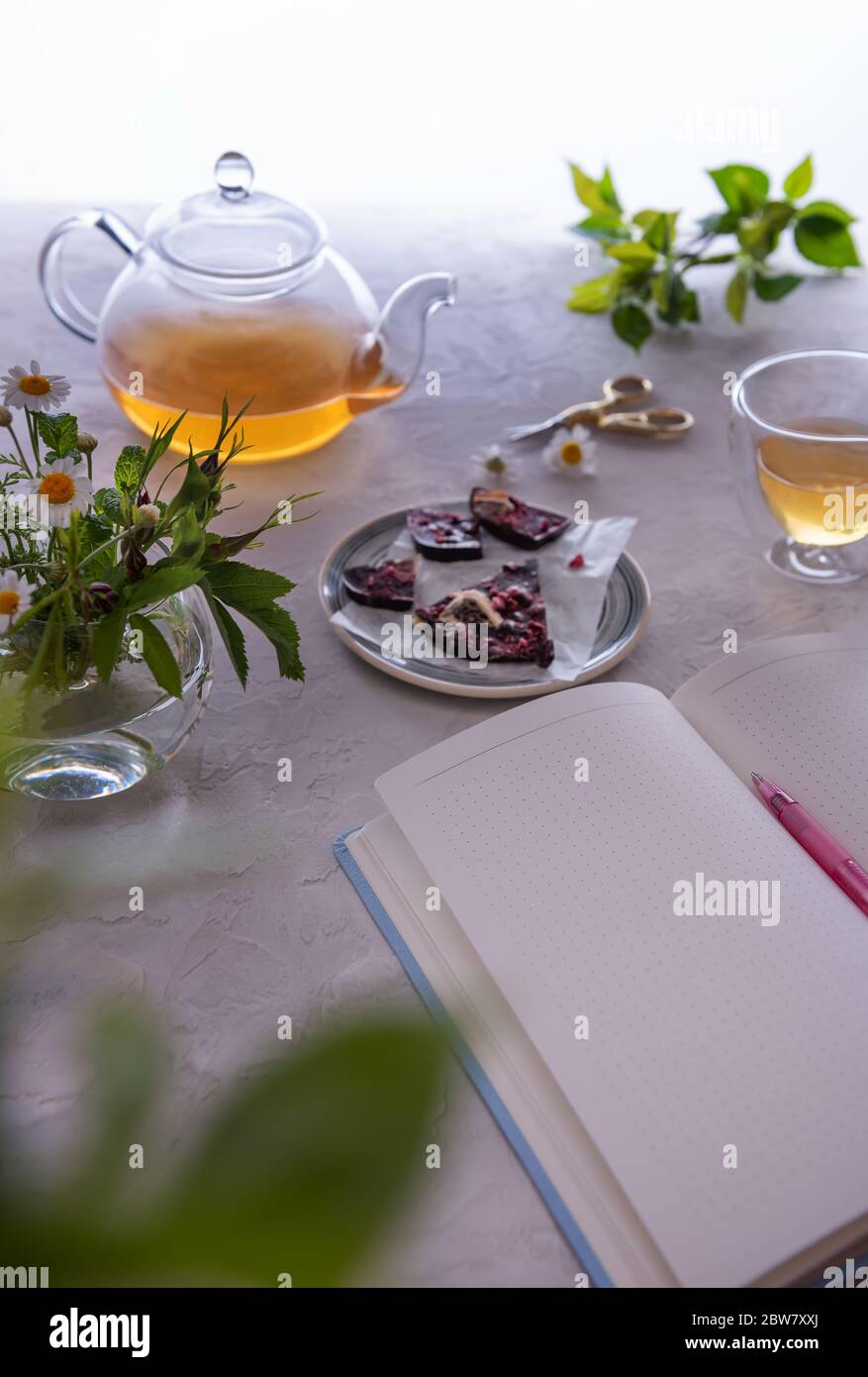 Journal ouvert à une page vide avec un stylo rose. Il y a aussi une théière en verre non concentrée, une tasse thermo avec thé, des chocolats et un bouquet de fleurs dans un vase. Banque D'Images