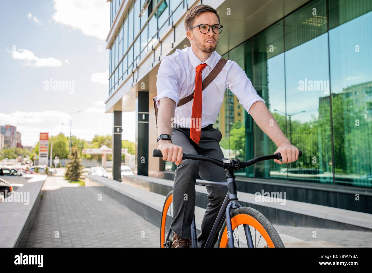 Beau jeune homme d'affaires en lunettes et vêtements de formaloport assis sur un vélo et se déplaçant le long de la route le long de l'architecture moderne Banque D'Images