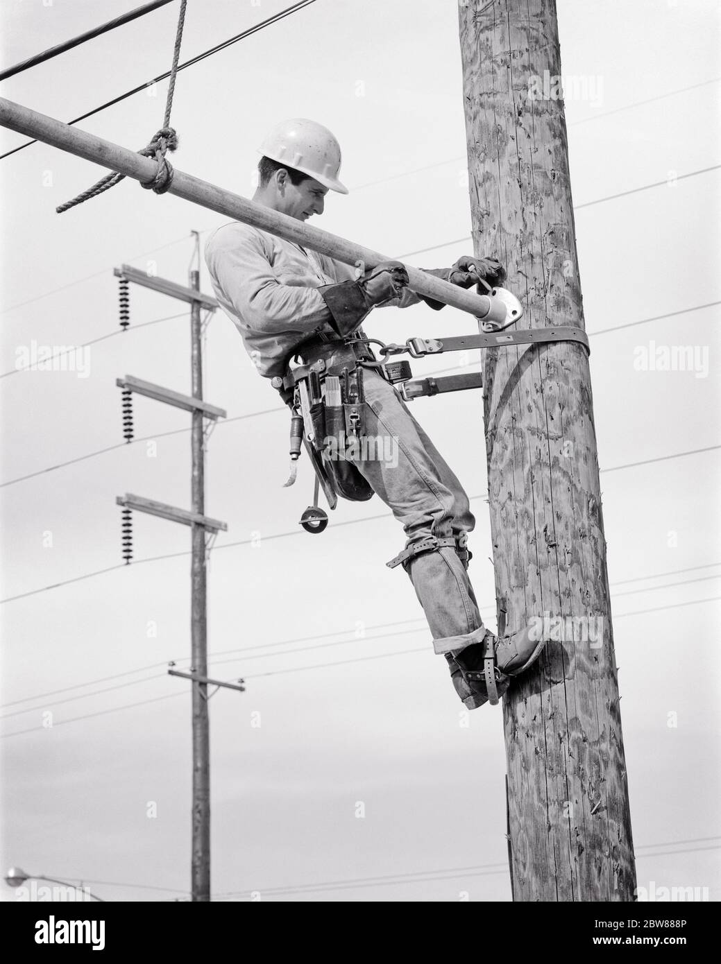 1960 LINEMAN ÉLECTRIQUE SUR POTEAU DE SERVICE FIXÉ AVEC CEINTURE DE SÉCURITÉ ET GAFFS OU DES ÉPERONS SUR SES PIEDS PORTANT UN CASQUE ET UNE CEINTURE À OUTILS - I3838 HAR001 HARS BLUE COLLIER COMPÉTENCES PROFESSIONNELLES COMPÉTENCES SON MANUEL ET ANGLE TRAVAIL EMPLOIS EMPLOYÉS DE SERVICE OU DE COMMERCE BLEU JEANS MI-ADULTE HOMME PRÉCISION SÉCURISÉ SPIRES NOIR ET BLANC DE LA GRILLE ETHNIQUE CAUCASIENNE HAR001 INSTALLATION LABIING DES SERVICES PUBLICS À L'ANCIENNE Banque D'Images