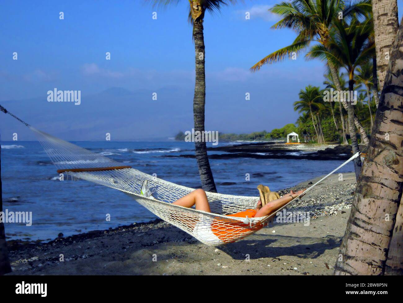 Les vacanciers se détend dans un hamac sur la côte de Kohala. La vue inclut les montagnes Mauna Kea et la plage de la baie de Makaiwa. Elle porte un chapeau de paille, orange Banque D'Images