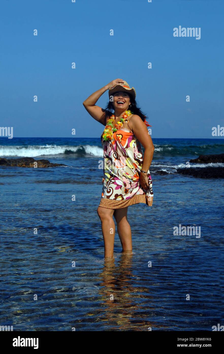 Belle femme se tient cheville profondément dans les eaux peu profondes de la Grande île d'Hawaï. Elle porte un maillot de bain aux couleurs vives, un chapeau de paille et un Banque D'Images