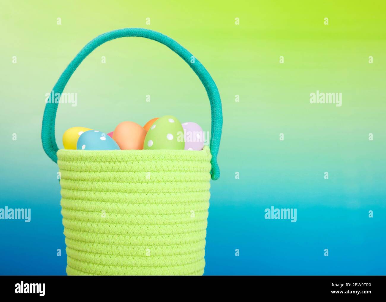 Gros plan sur un panier de Pâques tissé en fil vert vif avec poignée bleu clair rempli d'œufs de couleur festive avec des points, fond bleu dégradé à Banque D'Images