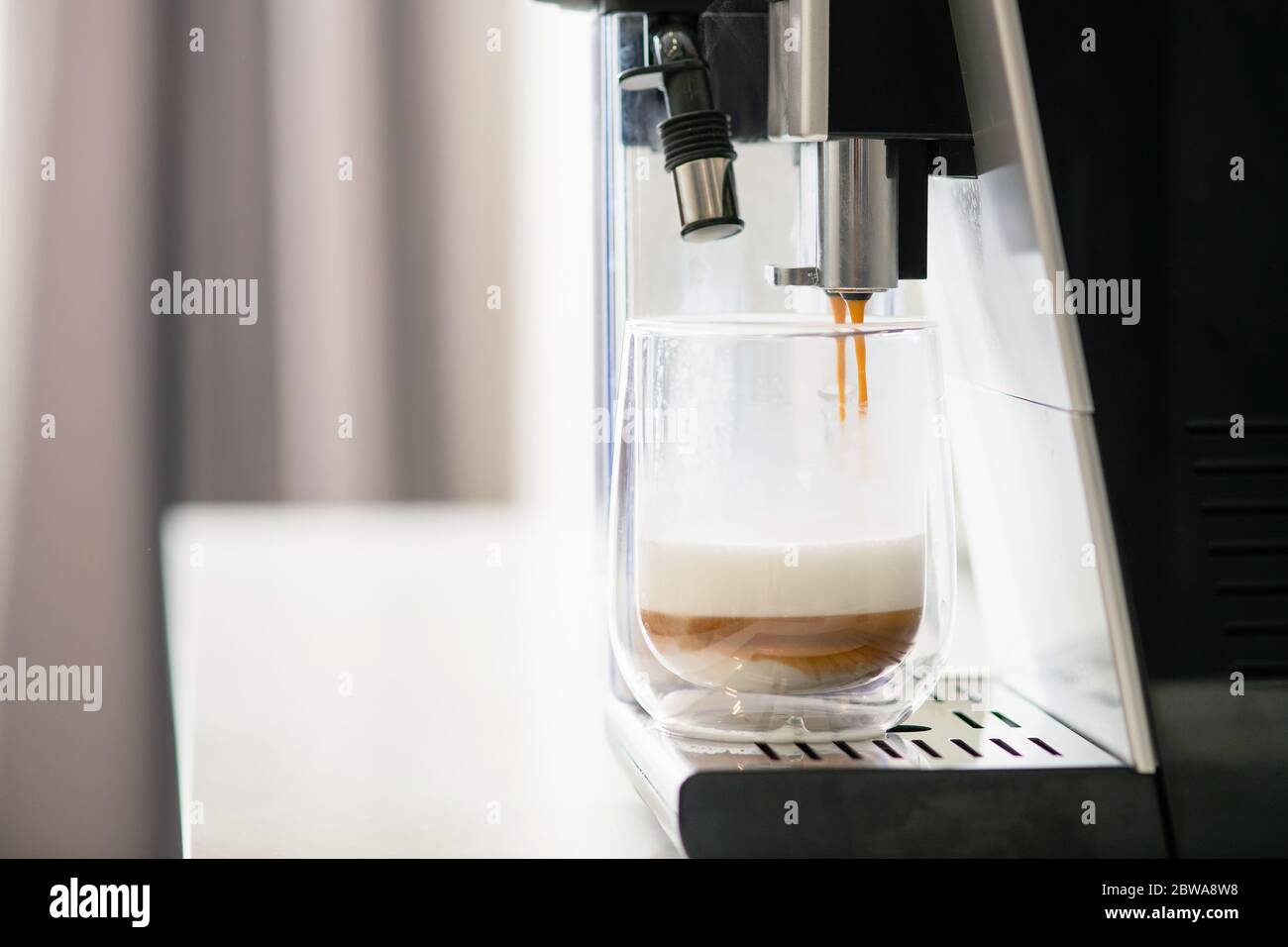 Préparation du café et du lait avec la machine à café, les liquides s'écoulent du filtre du support vers le verre transparent tasse par couches, préparation automatique de boissons chaudes au hom Banque D'Images