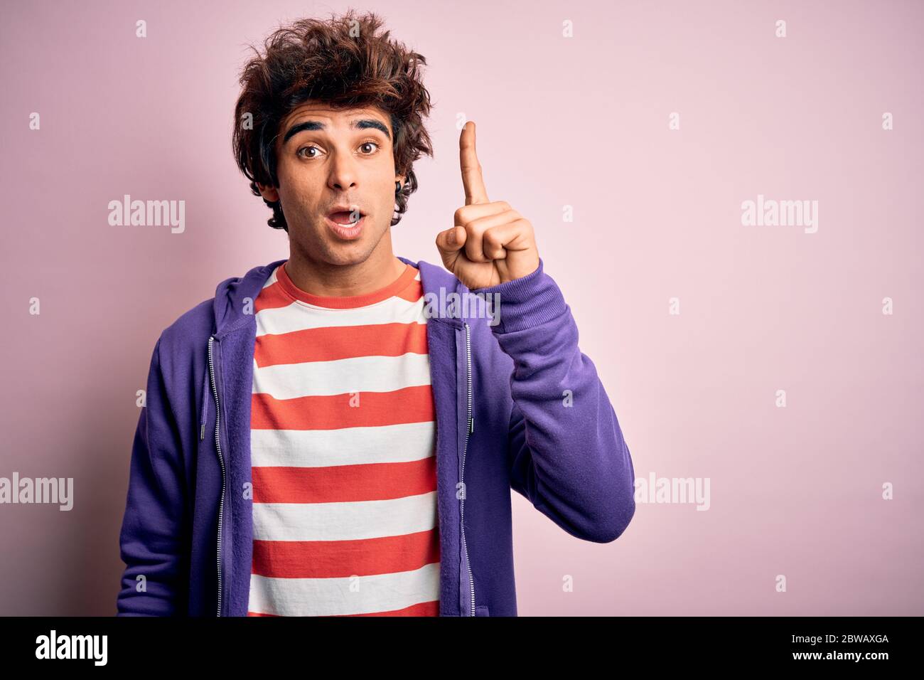 Jeune homme portant un t-shirt rayé et un sweat-shirt violet sur un arrière-plan rose isolé pointant le doigt vers le haut avec une idée réussie. Quitté et heureux. Numéro Banque D'Images
