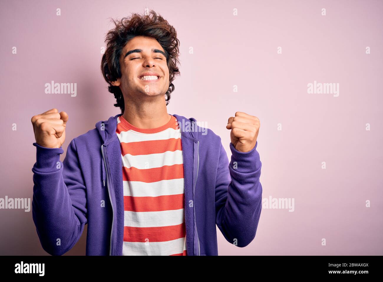 Jeune homme portant un t-shirt rayé et un sweat-shirt violet sur fond rose isolé très heureux et excité faisant le geste gagnant avec les bras levés, smi Banque D'Images