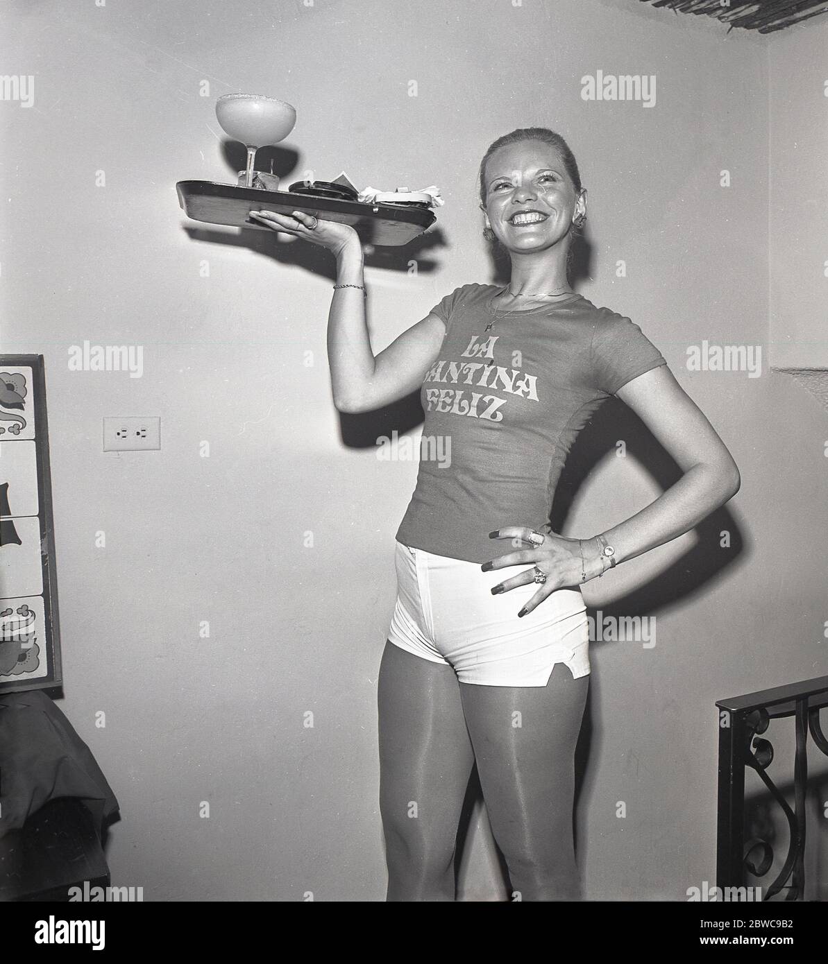 Années 1960, historique, une grande serveuse de cocktail mince en T-shirt et pantalon chaud donne un grand sourire portant un plateau de boissons dans un restaurant espagnol, la Santina Feliz, Los Angeles, Californie, Etats-Unis. Banque D'Images