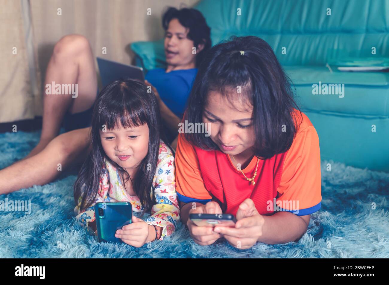 Famille dans le salon la nuit en utilisant un smartphone et une tablette chacun séparément, fille regardant la vidéo, père jouer au jeu et mère shopping Banque D'Images