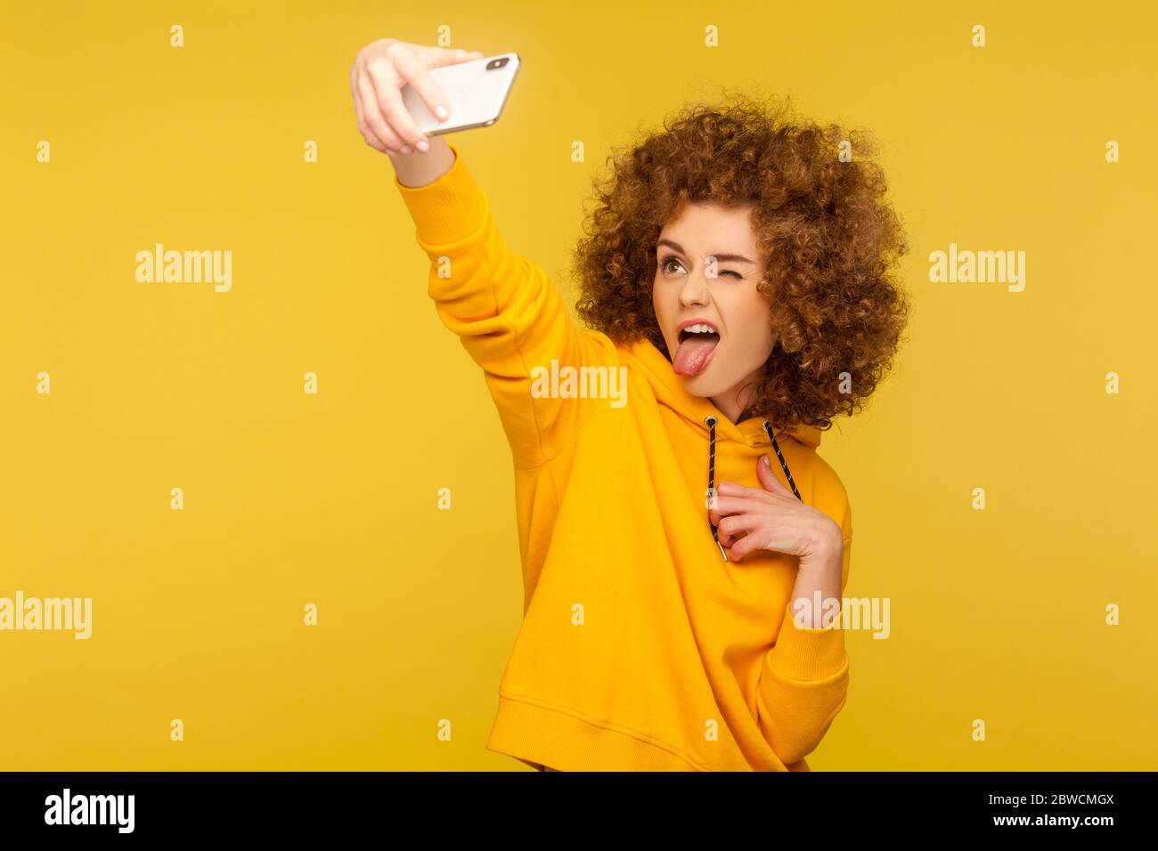 Portrait de la femme à cheveux bouclés sans souci dans un sweat à capuche de style urbain prenant le selfie et grimaquant avec la langue dehors, s'amuser tout en faisant la photo, vlog br Banque D'Images