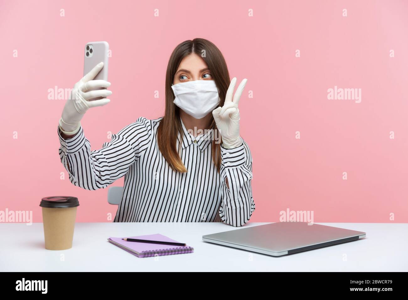 Femme optimiste assise en sécurité saine avec masque de protection et des gants au travail, montrant le geste de victoire au téléphone mobile, ayant en ligne communicati Banque D'Images