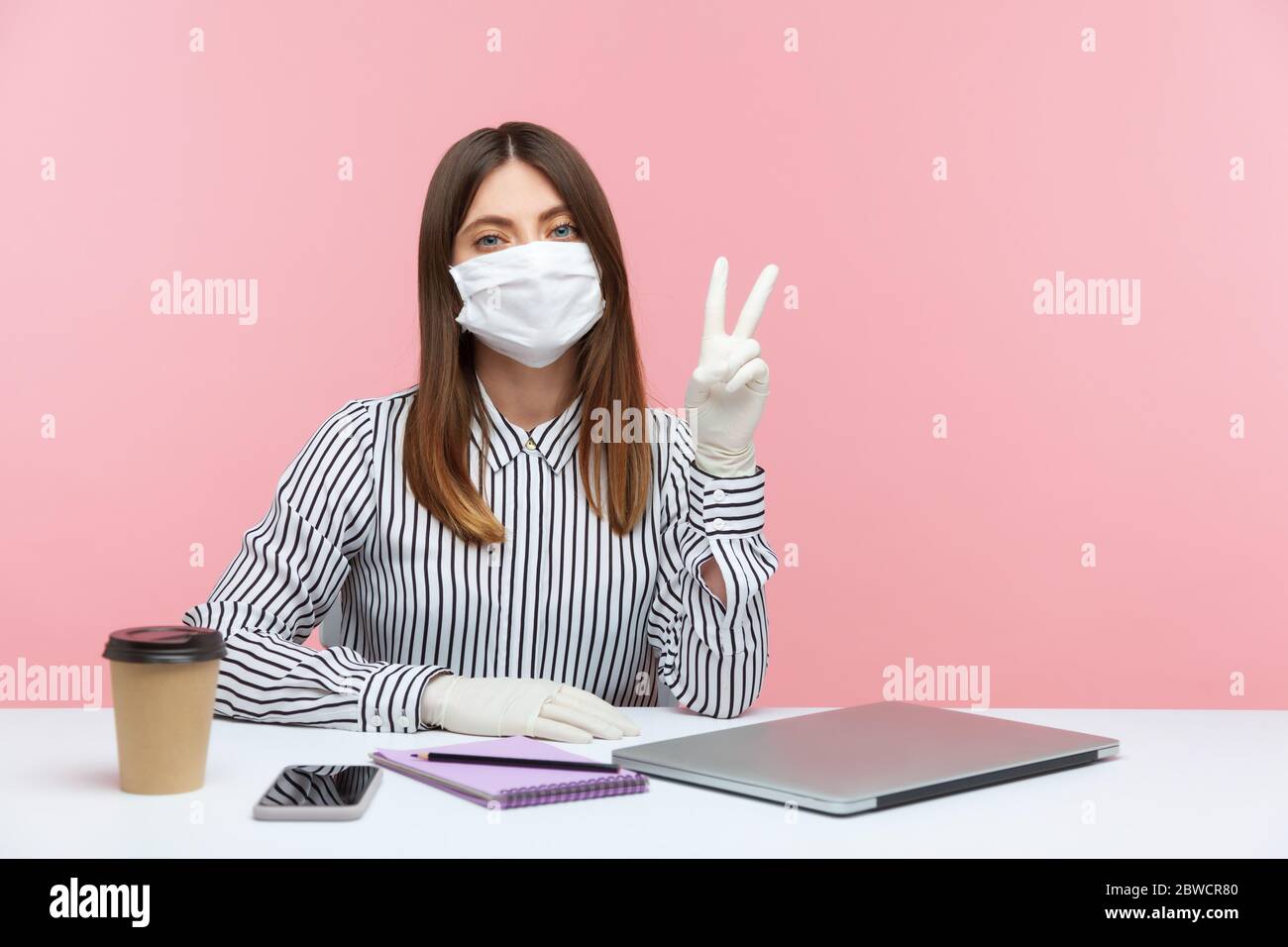 Femme optimiste employée assise en sécurité saine avec masque de protection et des gants pendant la quarantaine, montrant la victoire, geste de paix. Travailler chez soi Banque D'Images