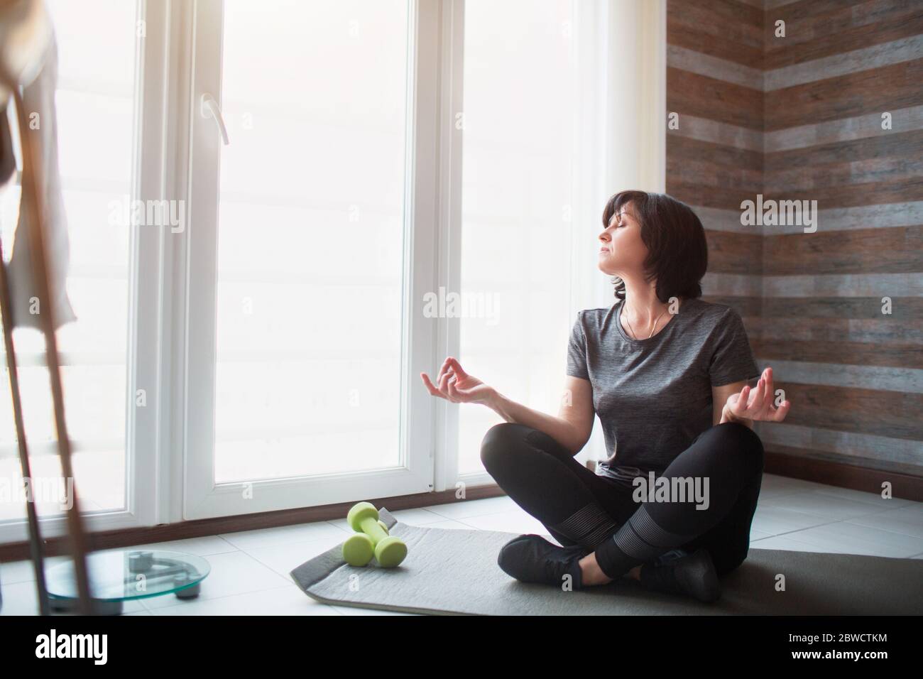 La femme fine pour adulte s'entraîner à la maison. Processus de méditation du modèle senior. Tenez les mains sur les genoux et regardez la fenêtre. Banque D'Images