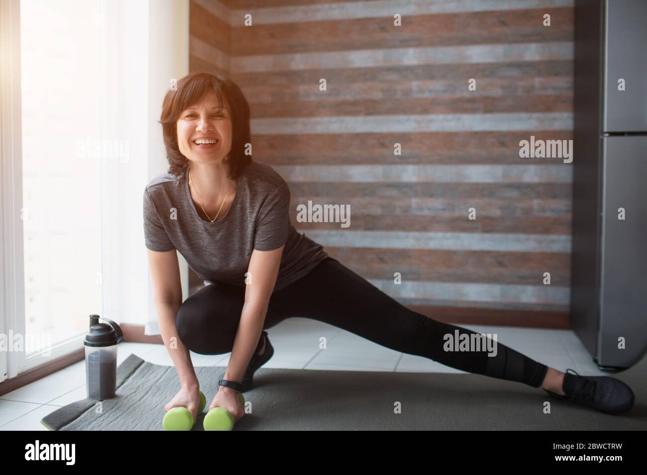 La femme fine pour adulte s'entraîner à la maison. Joyeux positif personne mature qui étire un côté de jambe et tient des haltères dans les mains. Entraînement à Banque D'Images