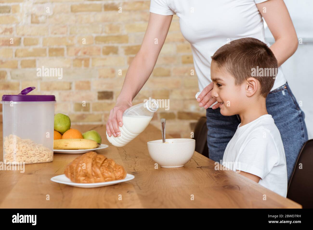 Mère versant du lait dans un bol avec des céréales près du fils souriant à la table de cuisine Banque D'Images